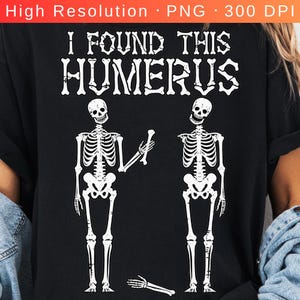 May include: A black t-shirt featuring a white graphic of two skeletons. One skeleton holds a bone, while the other stands nearby with a detached hand on the ground. The text "I FOUND THIS HUMERUS" is printed in a bone-shaped font above the skeletons.