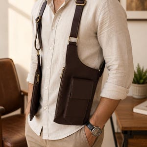 May include: A dark brown dual-pouch chest harness bag featuring adjustable straps with brass-toned metal hardware. The bag consists of two vertical rectangular pouches with zippered closures and a front flap pocket, worn over a light beige button-down shirt.