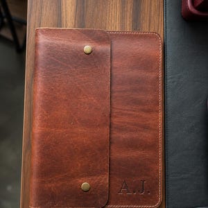 May include: A rectangular brown leather portfolio case featuring a wrap-around flap secured by two brass-colored metal snap buttons. The bottom right corner of the leather cover is embossed with the capital letters A.J. with visible stitching.