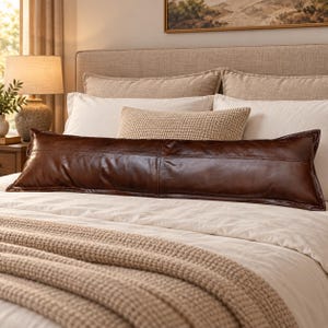 May include: A long, rectangular bolster pillow crafted from smooth, dark brown leather. The pillow features a horizontal seam running across the center and is finished with stitched edges, providing a structured and elongated shape for bedding.