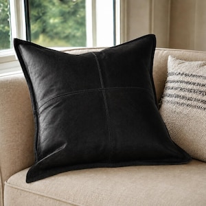May include: A square black leather throw pillow featuring a cross-stitched seam design that divides the front surface into four equal quadrants. The pillow has a structured edge and a smooth, matte finish, positioned upright on a light-colored sofa.