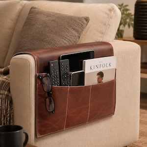 May include: A brown leather sofa armrest organizer featuring multiple pockets draped over the side of a beige couch. The organizer holds two black remote controls, a pair of black-rimmed eyeglasses, a smartphone, and a magazine titled KINFOLK.