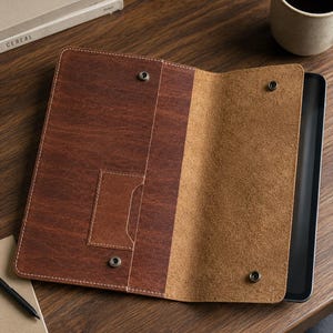 May include: A brown leather tablet sleeve featuring a smooth exterior, a textured interior lining, and four metal snap fasteners. The sleeve includes a rectangular pocket on the left side and is designed to hold a slim electronic tablet device securely.