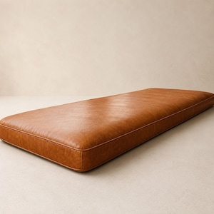 May include: A rectangular bench cushion upholstered in a smooth, tan-colored material with a visible grain texture. The cushion features clean, piped edges along the perimeter, creating a structured and uniform shape with a flat top surface.