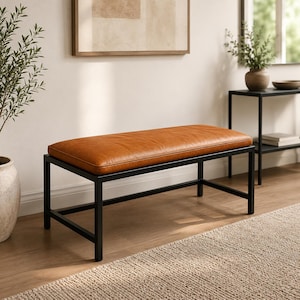 May include: A rectangular bench featuring a smooth, tan-colored upholstered seat cushion supported by a sturdy, black-painted metal frame with four straight legs and horizontal support bars connecting the base for structural stability.