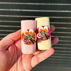 May include: Two cylindrical lighters, one light pink and one cream, each featuring a miniature three-dimensional bowl of ramen attached to the front. The ramen bowls contain noodles, sliced eggs, and pink and white fish cake slices.