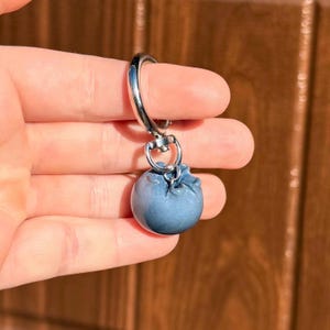 Può includere: Un portachiavi a forma di mirtillo con un fermaglio in metallo color argento. Il ciondolo è di colore blu tenue e ha una superficie strutturata. Il portachiavi è tenuto in mano su uno sfondo di legno sfocato.