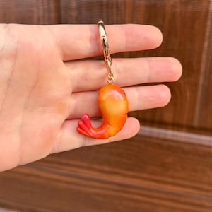 以下が含まれることがあります： ロブスタークラスプが付いたゴールドカラーのキーチェーンで、ミニチュアのエビのチャームが付いています。エビはオレンジと黄色で、赤い尾が付いており、気まぐれなアクセサリーです。