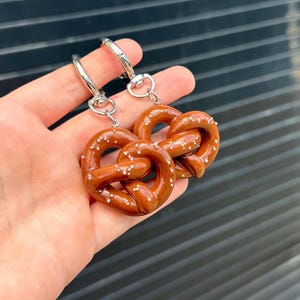 Puede incluir: Dos dijes de pretzel de color marrón con motas blancas que parecen sal, unidos a cierres de langosta de metal plateado. Los pretzels tienen un acabado brillante y liso con una forma anudada clásica, cada uno colgado de un anillo.