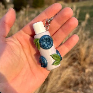 May include: A light purple lip balm tube with a white cap, decorated with blue flower and leaf accents. The tube is attached to a silver keychain clip. The design features a floral theme.