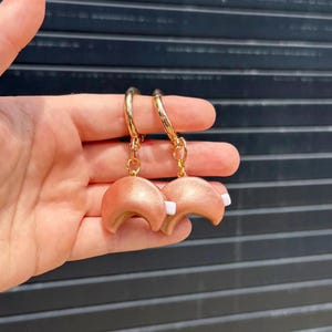 May include: Two metallic copper-coloured fortune cookie charms attached to gold-toned lobster claw clasps. Each crescent-shaped charm features a small white rectangular paper strip protruding from the centre, suspended by a gold jump ring.