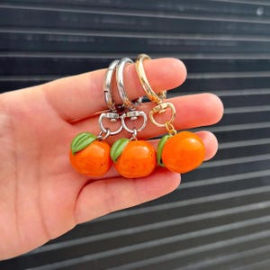 May include: Three orange fruit-shaped keychains with green leaves. Each keychain has a different metal ring: silver, silver, and gold. The keychains are held up against a dark background.