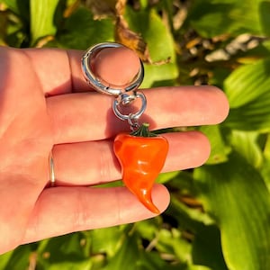 May include: A silver-colored keychain with a bright orange chili pepper charm. The pepper has a green stem and a curved shape. The keychain is held against a background of green foliage.