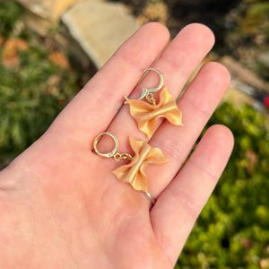 以下が含まれることがあります： 金色の蝶ネクタイ型のイヤリングと金色のフープのペア。イヤリングは、ぼやけた緑色の背景に対して、手で持たれています。
