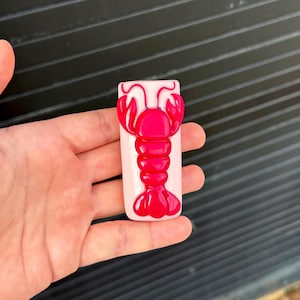 Pode incluir: Um objeto retangular rosa claro com uma figura de lagosta vermelha brilhante em relevo centrada na parte frontal. O design da lagosta inclui pinças detalhadas, um corpo segmentado e uma cauda em forma de leque, com acabamento liso.