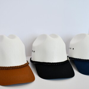 May include: Three white cowboy hats with different colored brims. The hats have a black braided band around the crown. The brims are brown, black, and navy blue.