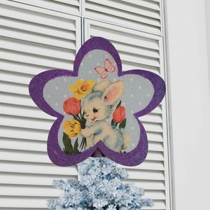 May include: Purple glitter star-shaped Easter decoration with a blue background featuring a cartoon bunny holding yellow and pink flowers. The bunny is surrounded by white polka dots.