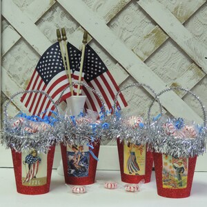 May include: Four red, glittery, mini buckets with handles, filled with candy and decorated with patriotic themed paper. The buckets are surrounded by silver tinsel and there are three American flags in the background.