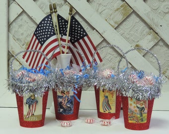 May include: Four red, glittery, mini buckets with handles, filled with candy and decorated with patriotic themed paper. The buckets are surrounded by silver tinsel and there are three American flags in the background.
