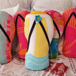A collection of colorful beach towel pillows shaped like flip-flops. The pillows feature various designs, including stripes, floral patterns, and a blue flower accent. The flip-flops have black straps and are arranged on a bed.