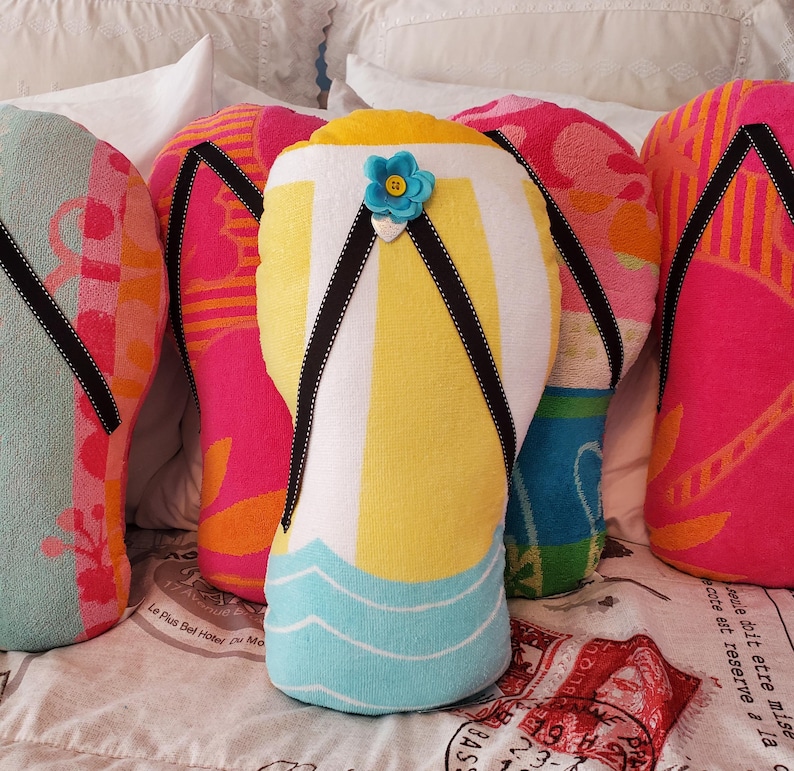 A collection of colorful beach towel pillows shaped like flip-flops. The pillows feature various designs, including stripes, floral patterns, and a blue flower accent. The flip-flops have black straps and are arranged on a bed.