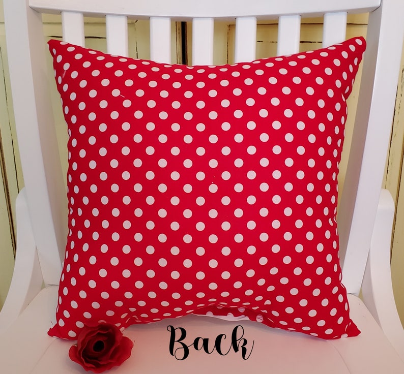 May include: Red pillow with white polka dots. The back of the pillow is shown. The pillow is sitting on a white chair.