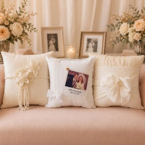 Peut inclure: Trois coussins décoratifs ivoire sur un canapé rose. L'un des coussins présente des ornements floraux et des glands, un autre une photo encadrée et le troisième un grand nœud. Deux photos encadrées et des compositions florales sont en arrière-plan.