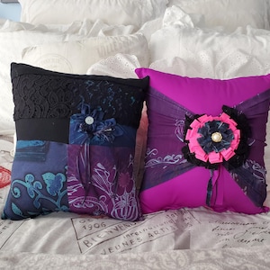 Two decorative memory throw pillows. One pillow is a patchwork of blue and purple fabrics with a black lace top and a fabric flower. The other is magenta with a large fabric flower and black accents. Both pillows are on a bed.