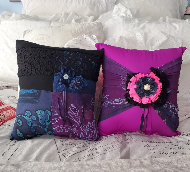 Two decorative memory throw pillows. One pillow is a patchwork of blue and purple fabrics with a black lace top and a fabric flower. The other is magenta with a large fabric flower and black accents. Both pillows are on a bed.