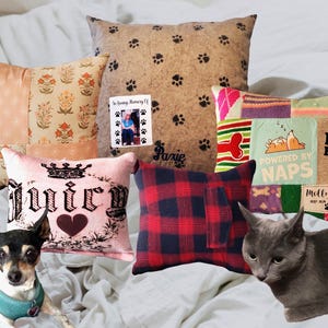 May include: A collection of decorative throw cushions featuring various patterns, including a tan paw print design with a memorial photo, a pink cushion with a crown and heart, a red and black plaid cushion, and a colourful patchwork design.