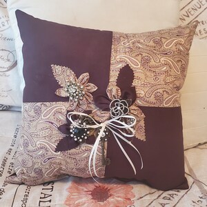 Square decorative memory pillow with a patchwork design. The pillow features a deep purple and paisley pattern fabric. Embellishments include fabric flowers, brooches, a key, and white ribbon. The pillow is approximately 14 inches square.