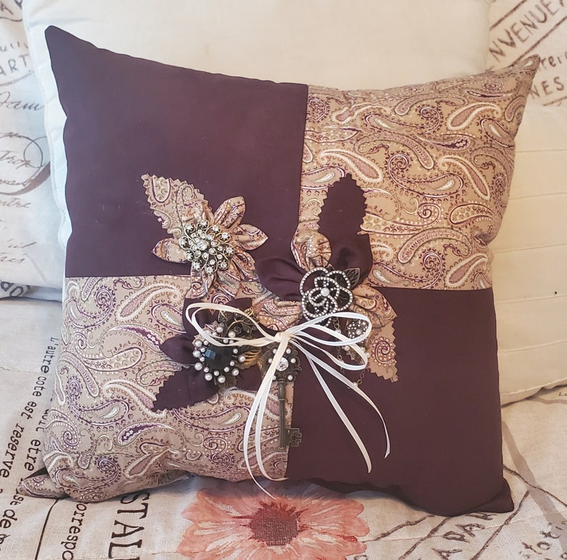 Square decorative memory pillow with a patchwork design. The pillow features a deep purple and paisley pattern fabric. Embellishments include fabric flowers, brooches, a key, and white ribbon. The pillow is approximately 14 inches square.