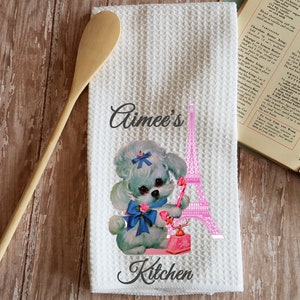 May include: A white kitchen towel with a pink and white Eiffel Tower, a blue and white poodle with a blue bow, and a pink rotary phone. The text "Aimee's Kitchen" is printed on the towel.