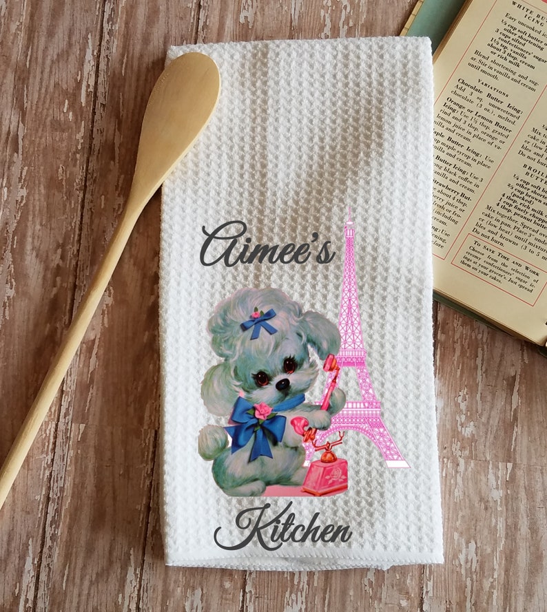 May include: A white kitchen towel with a pink and white Eiffel Tower, a blue and white poodle with a blue bow, and a pink rotary phone. The text "Aimee's Kitchen" is printed on the towel.
