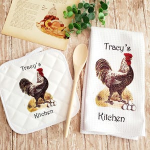 Puede incluir: Un salvamanteles acolchado blanco y un paño de cocina blanco con una ilustración de gallo. El texto "Tracy's Kitchen" está impreso en ambos artículos. Una cuchara de madera y una receta vintage también están en la imagen.