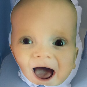 May include: A white pillow with a printed image of a baby's face. The baby is smiling with its mouth open and tongue sticking out. The pillow is soft and plush.