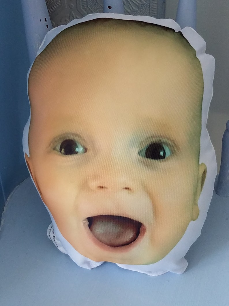 May include: A white pillow with a printed image of a baby's face. The baby is smiling with its mouth open and tongue sticking out. The pillow is soft and plush.