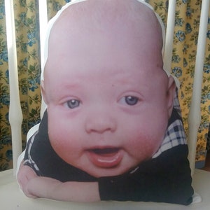 May include: A white pillow with a printed image of a baby wearing a black and white checkered shirt. The baby is smiling and looking at the camera.