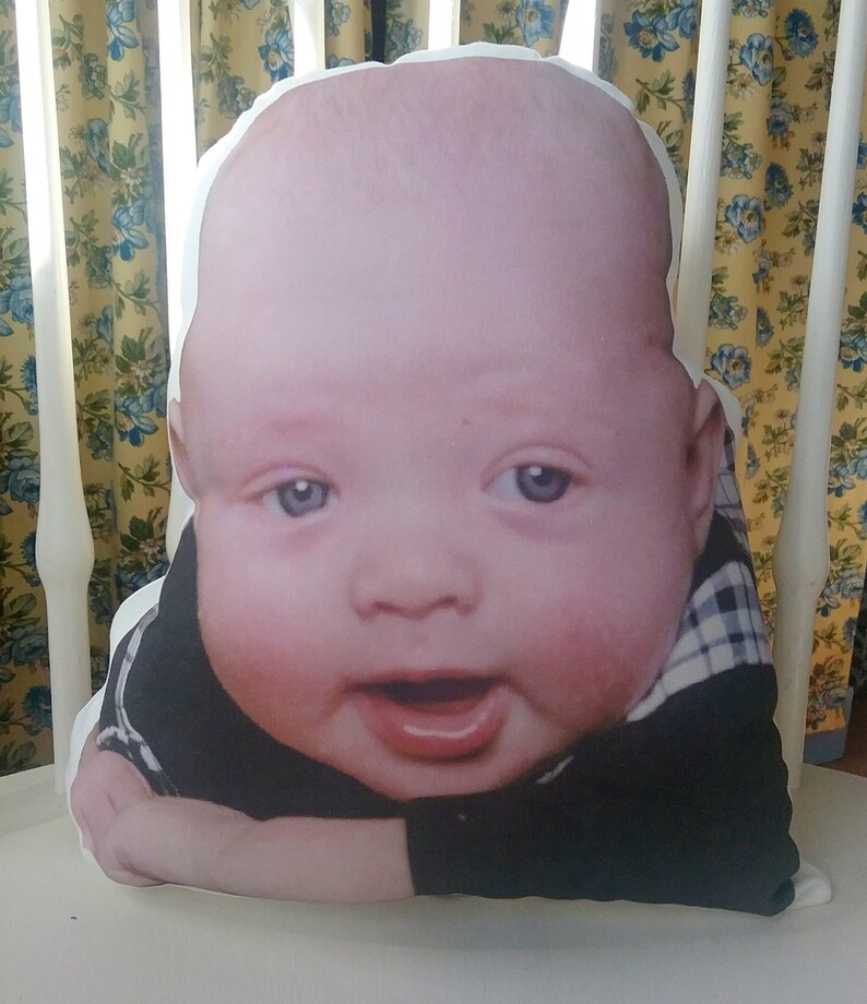 May include: A white pillow with a printed image of a baby wearing a black and white checkered shirt. The baby is smiling and looking at the camera.