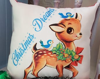 May include: White square pillow with a vintage illustration of a deer with a holly wreath and a red bow. Two blue birds perch on the deer. The words "Christmas Dreams" are written in blue script on the pillow.