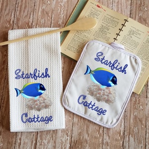 May include: White kitchen towel and pot holder with a blue and yellow fish and the text "Starfish Cottage" in a blue rope design.