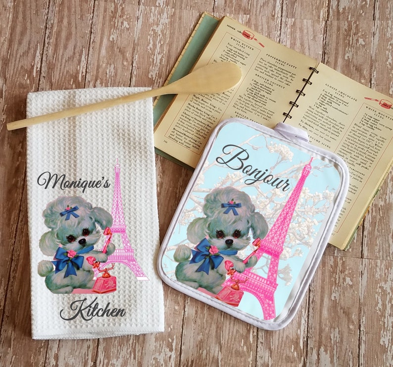 May include: A white kitchen towel with a pink Eiffel Tower and a poodle holding a pink phone. The text "Monique's Kitchen" is printed on the towel.