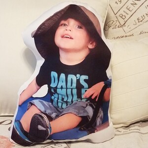 May include: A white pillow with a printed image of a young child wearing a brown hat and a black t-shirt that says "Dad's Smile". The child is sitting on a blue surface and looking up.