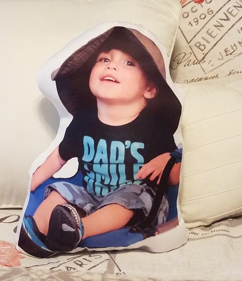 May include: A white pillow with a printed image of a young child wearing a brown hat and a black t-shirt that says "Dad's Smile". The child is sitting on a blue surface and looking up.