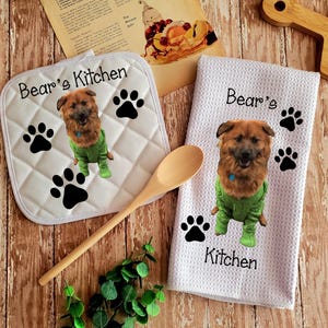May include: White kitchen towel and pot holder with a brown dog wearing a green sweater and paw prints. The towel says "Bear's Kitchen" and the pot holder says "Bear's Kitchen".