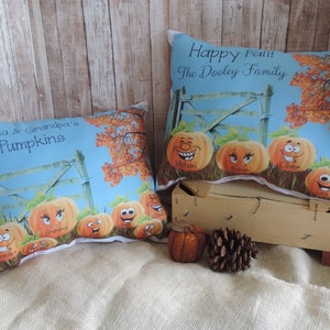Könnte beinhalten: Zwei dekorative Kissen mit Herbstthema. Auf einem Kissen steht "Grandma & Grandpa's Pumpkins" und auf dem anderen "Happy Fall! The Dooley Family". Beide Kissen zeigen Cartoon-Kürbisse mit Gesichtern und einem blauen Hintergrund.