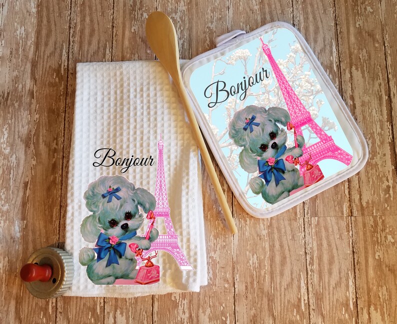 May include: A white kitchen towel and a matching pot holder with a blue and white poodle holding a pink phone in front of the Eiffel Tower. The text "Bonjour" is printed on both items.