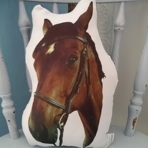 Pu&ograve; includere: Un cuscino bianco con la testa di un cavallo marrone stampata su di esso. Il cavallo indossa una briglia e ha una stella bianca sulla fronte.