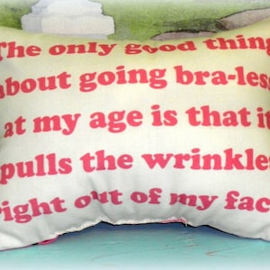 May include: A white pillow with pink text that reads "The only good thing about going bra-less at my age is that it pulls the wrinkles right out of my face."