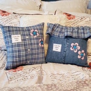 Two decorative pillows made from clothing. One is made from a blue plaid shirt, the other from denim. Both have appliqued pink flowers with blue centers. The plaid pillow has the text "I Love You Emma - Dad". The denim pillow has the text "I Love You Sydney - Dad".